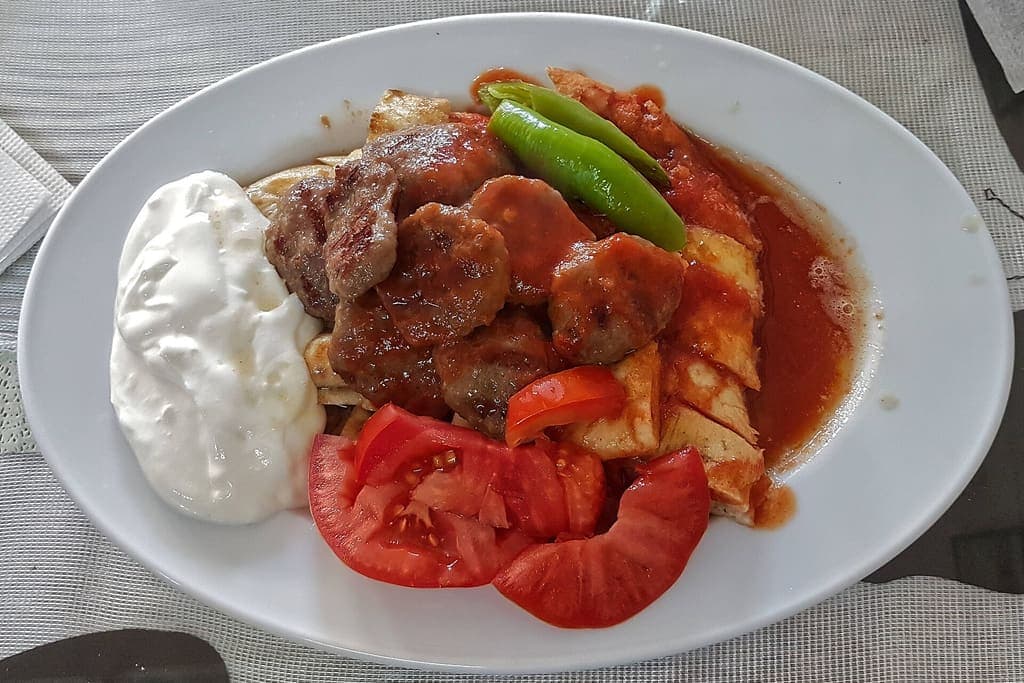 Beyaz tabakta pide üstünde köfte, domates sosu, yoğurt, domates ve yeşil biber